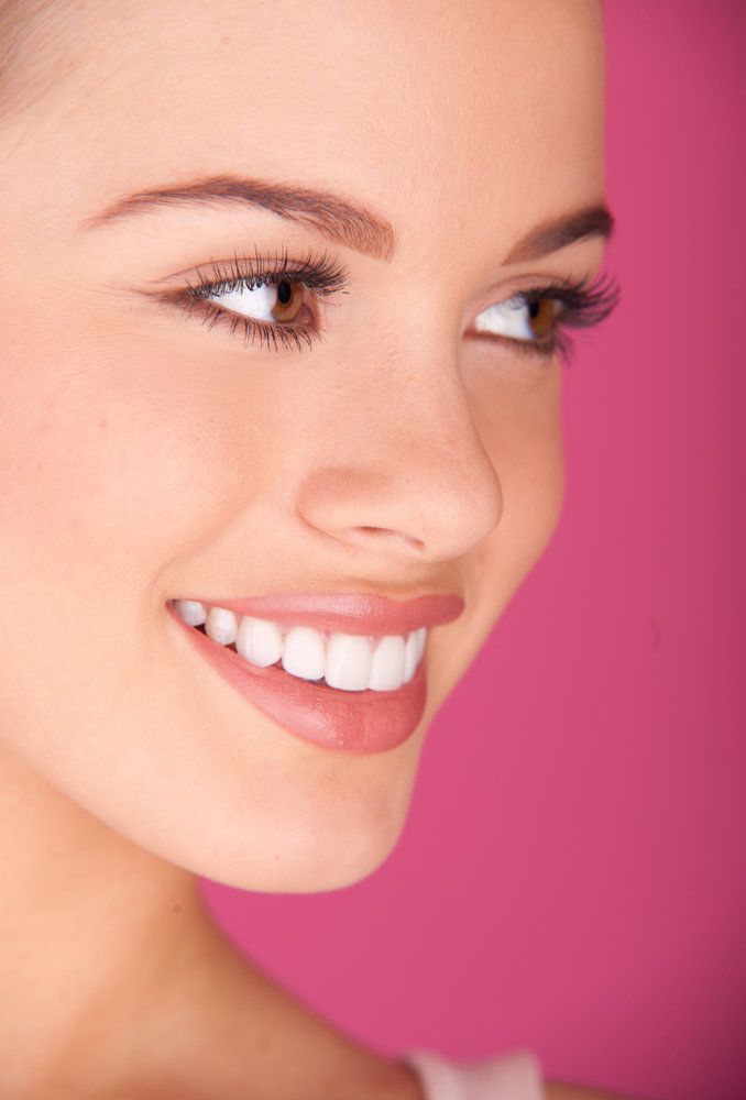 A beautiful, young woman with brown eyes smiles in front of a pink background.
